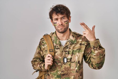 Foto de Joven hispano vistiendo uniforme camuflaje del ejército disparando y matándose apuntando de la mano y los dedos a la cabeza como arma, gesto suicida. - Imagen libre de derechos