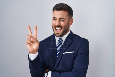 Téléchargez les photos : Un bel homme hispanique portant costume et cravate souriant avec un clin d'œil joyeux à la caméra faisant signe de victoire avec les doigts. numéro deux. - en image libre de droit