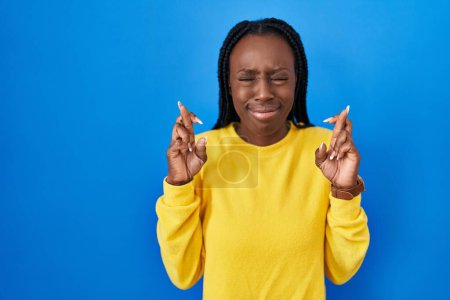 Foto de Beautiful black woman standing over blue background gesturing finger crossed smiling with hope and eyes closed. luck and superstitious concept. - Imagen libre de derechos