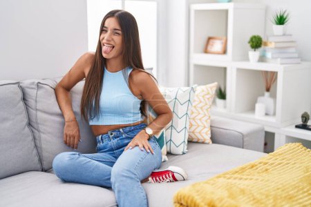 Photo for Young brunette woman sitting on the sofa at home sticking tongue out happy with funny expression. emotion concept. - Royalty Free Image