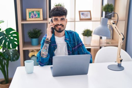 Téléchargez les photos : Jeune homme hispanique utilisant un ordinateur portable et parlant sur smartphone à la maison - en image libre de droit