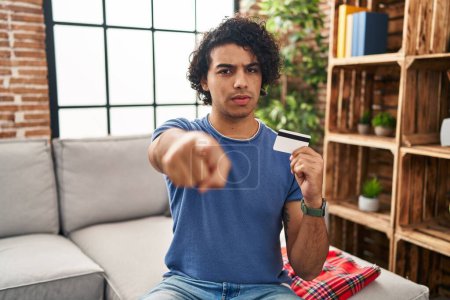 Photo for Hispanic man with curly hair holding credit card pointing with finger to the camera and to you, confident gesture looking serious - Royalty Free Image