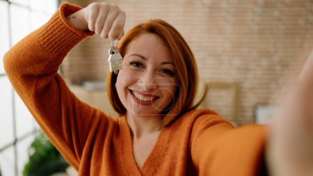 Photo for Young redhead woman making selfie by the camera holding key of new house at new home - Royalty Free Image