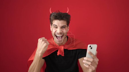 Téléchargez les photos : Jeune homme hispanique portant costume diable en utilisant smartphone sur fond rouge isolé - en image libre de droit