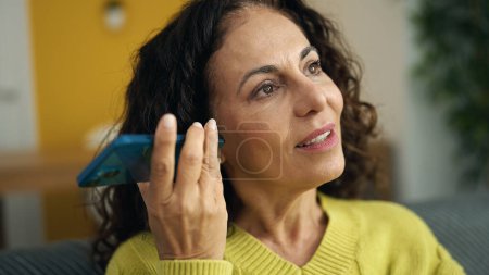 Photo for Middle age hispanic woman listening voice message by smartphone sitting on sofa at home - Royalty Free Image