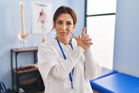Photo for Young brunette woman working at pain recovery clinic holding symbolic gun with hand gesture, playing killing shooting weapons, angry face - Royalty Free Image