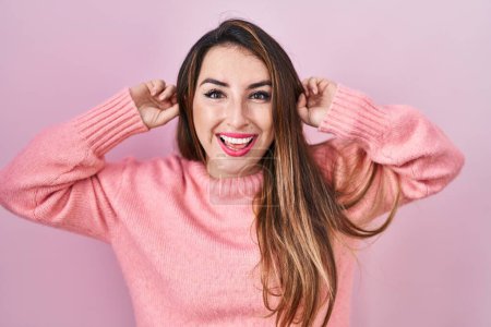 Foto de Mujer hispana joven de pie sobre fondo rosa sonriendo tirando de las orejas con los dedos, gesto divertido. problema de audición - Imagen libre de derechos