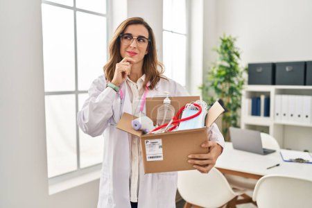 Photo for Young doctor woman holding box with medical items serious face thinking about question with hand on chin, thoughtful about confusing idea - Royalty Free Image