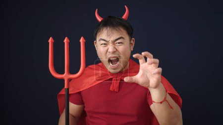 Téléchargez les photos : Jeune homme chinois portant costume diable faisant geste effrayant sur fond noir isolé - en image libre de droit
