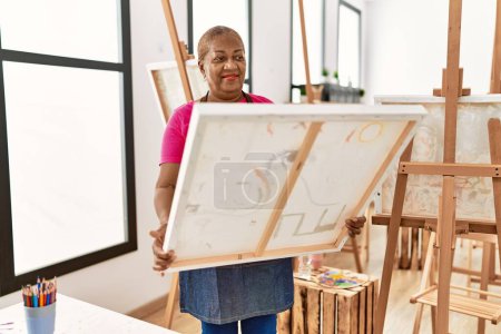 Foto de Senior africana americana mujer sonriendo confiado buscando dibujar lienzo en estudio de arte - Imagen libre de derechos