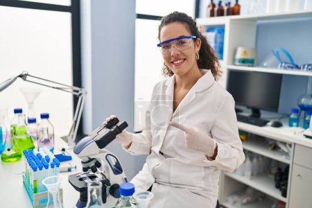 Téléchargez les photos : Jeune femme hispanique travaillant au laboratoire scientifique souriant heureux pointant avec la main et le doigt - en image libre de droit