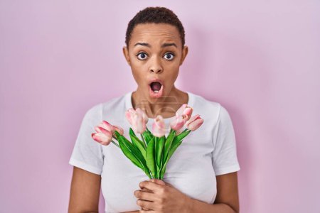 Téléchargez les photos : Belle femme afro-américaine tenant un bouquet de fleurs de tulipes roses effrayées et choquées par l'expression surprise et étonnante, la peur et le visage excité. - en image libre de droit
