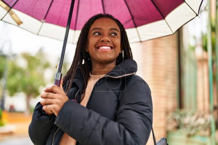 Téléchargez les photos : Afro-américaine femme souriant confiant en utilisant parapluie à la rue - en image libre de droit
