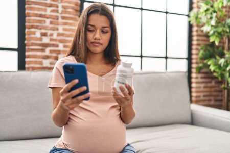 Téléchargez les photos : Jeune femme latine enceinte à l'aide d'un smartphone tenant bouteille de pilules à la maison - en image libre de droit