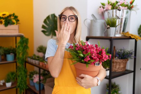 Foto de Mujer joven caucásica que trabaja en floristería sosteniendo la planta cubriendo la boca con la mano, conmocionada y temerosa de equivocarse. expresión sorprendida - Imagen libre de derechos