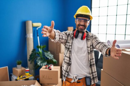 Téléchargez les photos : Jeune homme hispanique avec barbe travaillant à la rénovation de la maison regardant la caméra souriant à bras ouverts pour un câlin. expression joyeuse embrassant le bonheur. - en image libre de droit