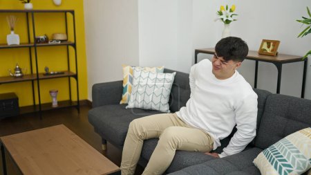Foto de Joven hispano sufriendo de dolor de espalda sentado en el sofá en casa - Imagen libre de derechos
