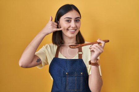Téléchargez les photos : Fille hispanique manger cuillère en bois sain souriant faire geste de téléphone avec la main et les doigts comme parler au téléphone. communiquer les concepts. - en image libre de droit