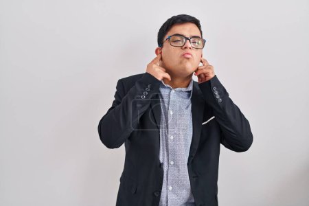 Foto de Joven hombre hispano con síndrome de Down con estilo de negocios cubriendo oídos con dedos con expresión molesta por el ruido de la música alta. concepto de sordo. - Imagen libre de derechos