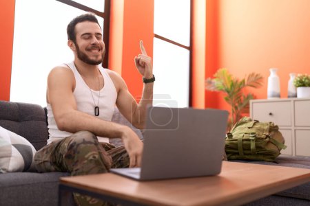 Foto de Joven hombre hispano usando uniforme camuflaje del ejército haciendo videollamadas en casa sonriendo con una idea o pregunta señalando con el dedo con cara feliz, número uno - Imagen libre de derechos