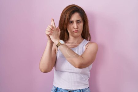 Téléchargez les photos : Femme brune debout sur fond rose tenant une arme symbolique avec geste de la main, jouant à tuer des armes à feu, visage en colère - en image libre de droit