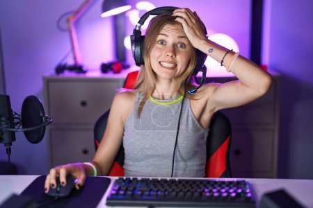 Foto de Mujer rubia caucásica jugando videojuegos con auriculares estresados y frustrados con la mano en la cabeza, cara sorprendida y enojada - Imagen libre de derechos