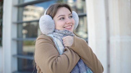 Téléchargez les photos : Jeune femme blonde souriante et confiante portant des cache-oreilles et une écharpe dans la rue - en image libre de droit