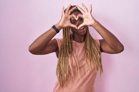 Photo for African american woman with braided hair standing over pink background doing heart shape with hand and fingers smiling looking through sign - Royalty Free Image