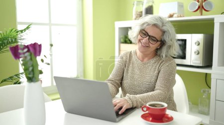 Foto de Mujer de mediana edad con cabello gris usando portátil sentado en la mesa en casa - Imagen libre de derechos