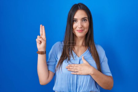 Téléchargez les photos : Young brunette woman standing over blue background smiling swearing with hand on chest and fingers up, making a loyalty promise oath - en image libre de droit