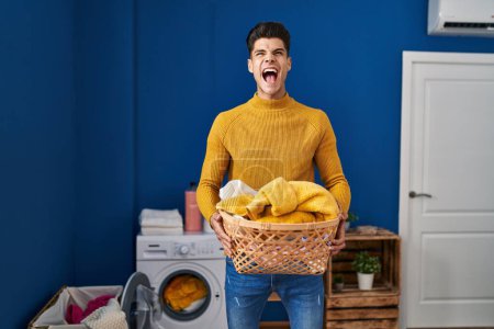 Photo for Young hispanic man holding laundry basket angry and mad screaming frustrated and furious, shouting with anger looking up. - Royalty Free Image