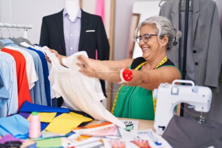 Téléchargez les photos : Femme aux cheveux gris d'âge moyen tailleur souriant confiant tenant t-shirt au magasin de tailleurs - en image libre de droit