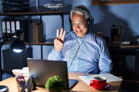 Photo for Hispanic senior man wearing call center agent headset at night showing and pointing up with fingers number three while smiling confident and happy. - Royalty Free Image