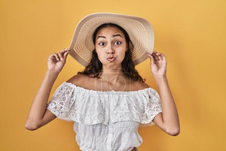 Téléchargez les photos : Jeune femme brésilienne portant un chapeau d'été sur fond jaune gonflant les joues avec un visage drôle. bouche gonflée d'air, attrapant l'air. - en image libre de droit