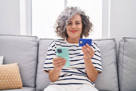 Téléchargez les photos : Femme aux cheveux gris du moyen âge utilisant un smartphone et une carte de crédit assise sur un canapé à la maison - en image libre de droit