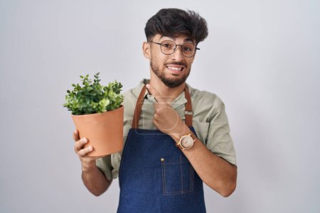 Téléchargez les photos : Homme arabe avec barbe tenant pot de plante verte pointant de côté inquiet et nerveux avec index, expression préoccupée et surprise - en image libre de droit