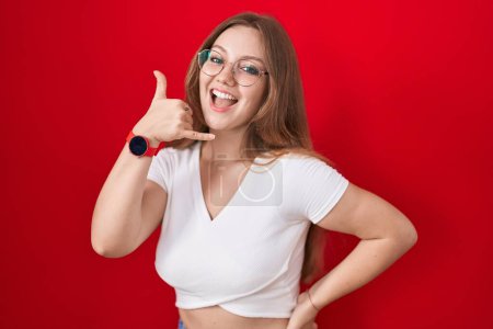 Téléchargez les photos : Jeune femme caucasienne debout sur fond rouge souriant faire geste de téléphone avec la main et les doigts comme parler au téléphone. communiquer les concepts. - en image libre de droit