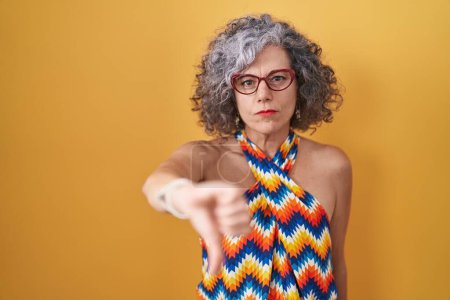 Photo for Middle age woman with grey hair standing over yellow background looking unhappy and angry showing rejection and negative with thumbs down gesture. bad expression. - Royalty Free Image