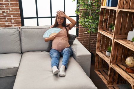 Foto de Mujer latina joven embarazada usando ventilador de mano sentado en el sofá en casa - Imagen libre de derechos