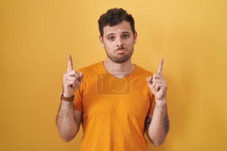 Young hispanic man standing over yellow background pointing up looking sad and upset, indicating direction with fingers, unhappy and depressed. 
