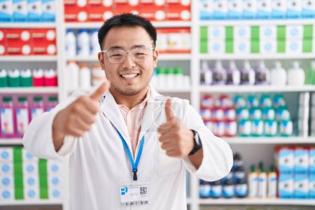 Foto de Joven chino trabajando en farmacia aprobando hacer gesto positivo con la mano, pulgares arriba sonriendo y feliz por el éxito. gesto ganador. - Imagen libre de derechos