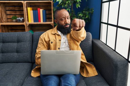 Téléchargez les photos : Afro-Américain utilisant un ordinateur portable à la maison assis sur le canapé pointant du doigt la caméra et à vous, geste confiant regardant sérieux - en image libre de droit