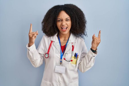 Photo for Young african american woman wearing doctor uniform and stethoscope shouting with crazy expression doing rock symbol with hands up. music star. heavy music concept. - Royalty Free Image