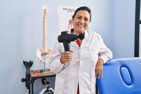 Photo for Middle age hispanic doctor woman holding therapy massage gun at wellness center looking positive and happy standing and smiling with a confident smile showing teeth - Royalty Free Image