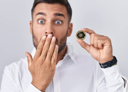 Foto de Hombre hispano guapo sosteniendo moneda criptomoneda cardano cubriendo la boca con la mano, sorprendido y asustado por error. expresión sorprendida - Imagen libre de derechos