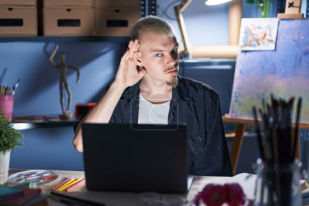 Téléchargez les photos : Jeune homme caucasien utilisant un ordinateur portable la nuit au studio d'art souriant avec la main sur l'oreille en écoutant une rumeur ou des commérages. concept de surdité. - en image libre de droit