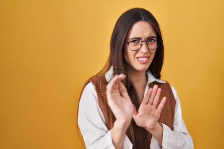 Photo for Young brunette woman standing over yellow background wearing glasses disgusted expression, displeased and fearful doing disgust face because aversion reaction. with hands raised - Royalty Free Image