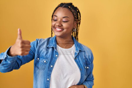 Téléchargez les photos : Femme afro-américaine avec des tresses debout sur fond jaune regardant fier, souriant faire les pouces vers le haut geste sur le côté - en image libre de droit
