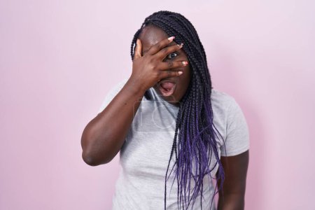 Foto de Joven mujer africana de pie sobre fondo rosa asomándose en shock cubriendo la cara y los ojos con la mano, mirando a través de los dedos con expresión avergonzada. - Imagen libre de derechos