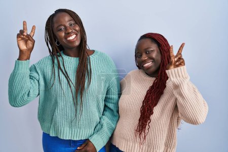 Téléchargez les photos : Deux femmes africaines debout sur fond bleu souriant regardant la caméra montrant des doigts faisant signe de victoire. numéro deux. - en image libre de droit